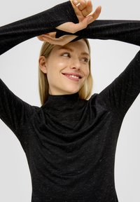 Chemise à col roulé noire à manches longues avec un design ajusté et un tissu texturé, présentant un subtil fil métallique et un décolleté drapé.