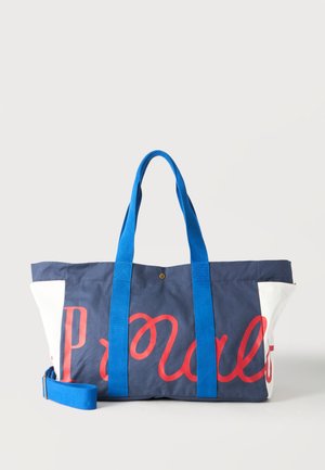 Sac cabas bleu marine et blanc avec des poignées bleu vif et un texte rouge en cursive sur le devant, sur un fond clair uni.