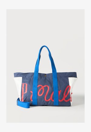 Sac cabas bleu marine et blanc avec des poignées bleu vif et un texte rouge en cursive sur le devant, sur un fond clair uni.