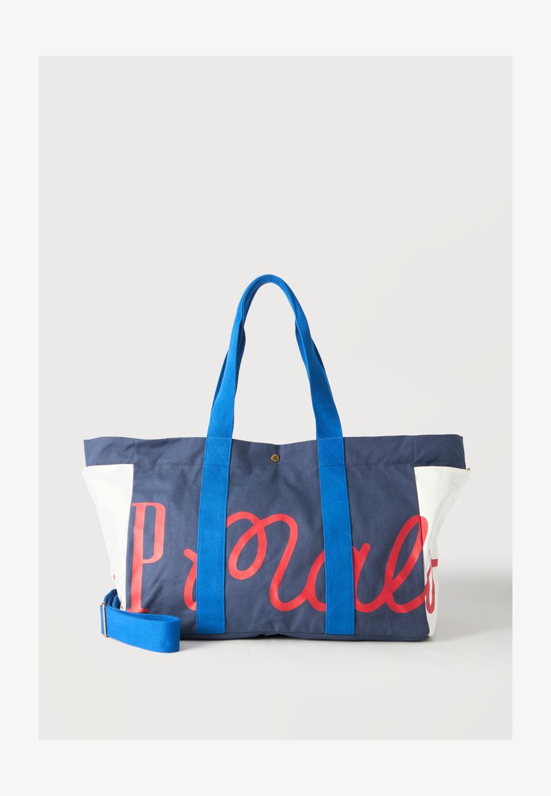 Sac cabas bleu marine et blanc avec des poignées bleu vif et un texte rouge en cursive sur le devant, sur un fond clair uni.