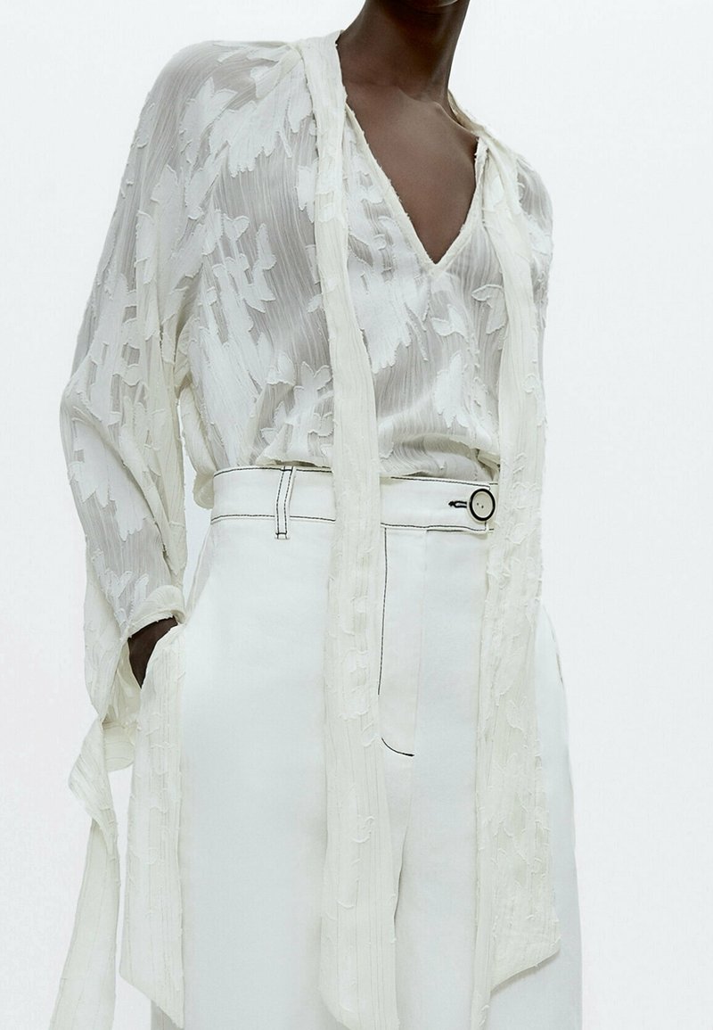 Personne à la peau foncée portant un chemisier blanc texturé avec de longues attaches et un pantalon blanc taille haute avec un bouton rond.