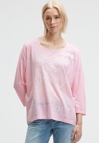 Blonde woman wearing a loose pink top with text "Reach your Dreams" and light blue jeans, standing against a plain background.