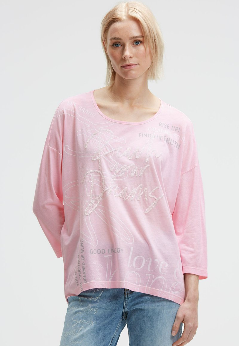 Blonde woman wearing a loose pink top with text "Reach your Dreams" and light blue jeans, standing against a plain background.