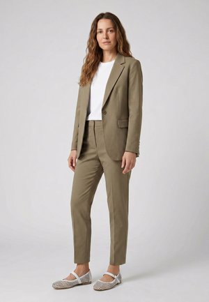 Femme aux longs cheveux ondulés, portant une veste et un pantalon de costume beige, un top blanc et des ballerines blanches à motifs Mary Jane, sur un fond uni.