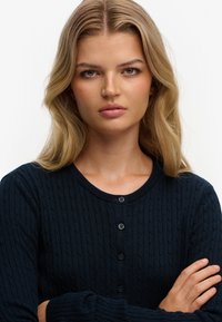 Marineblå cardigan med kabelstrik, rund halsudskæring, ribstrikkede manchetter og lukning med fem knapper. Teksturen er glat med et subtilt mønster.