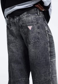 Jeans in denim grigio scuro con vestibilità rilassata. Caratteristiche includono una toppa con logo a triangolo, tasche posteriori e dettaglio verticale testurizzato sul lato.