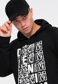Felpa nera con una stampa bianca audace "DENIM", cappuccio con lacci e cappellino da baseball nero. Presenta un design di tessuto pulito e texture.