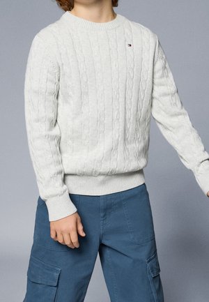 Barn iført en hvid kabelstrikket sweater med et lille logo og blå cargobukser mod en ensfarvet grå baggrund.