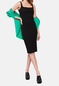 Robe noire moulante avec encolure carrée, portée avec une veste verte oversize. Le mannequin porte des chaussures à plateforme noires. Tissu lisse, silhouette ajustée.