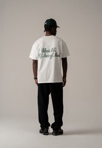 Hvit bomulls t-skjorte med grønn tekst "Blkvis O.C. NY State of Mind" på ryggen, kombinert med svarte bukser og mørke sko.