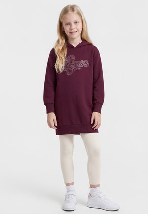 Blondynka stoi, ubrana w bordową sukienkę z kapturem z logo "Guess", kremowe legginsy, białe skarpetki i białe tenisówki.