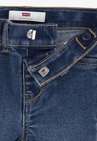 Jean en denim bleu foncé avec fermeture à bouton et clip en argent, accents de couture jaune et étiquette Levi's au niveau de la taille.
