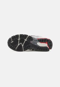 Semelle de chaussure de course noire et grise avec accents rouges, motif profond et rainures courbes pour adhérence et flexibilité sur fond blanc.