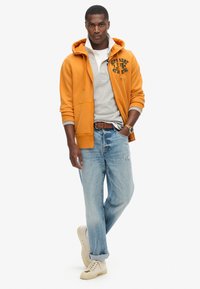 Felpa arancione con tasche frontali sopra una polo grigia, abbinata a jeans leggeri in denim strappati e sneakers beige. Orologio da polso visibile.