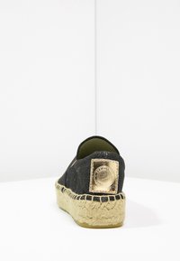Espadrille en tissu noir avec une surface texturée, semelle en jute et plaque en métal doré sur le talon. Design minimaliste, style à enfiler.