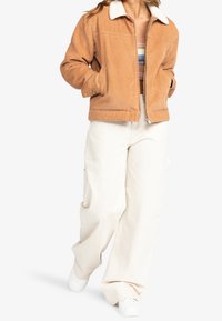 Veste en velours côtelé marron clair avec un col en fourrure blanche, associée à un pantalon ample crème et des baskets blanches.