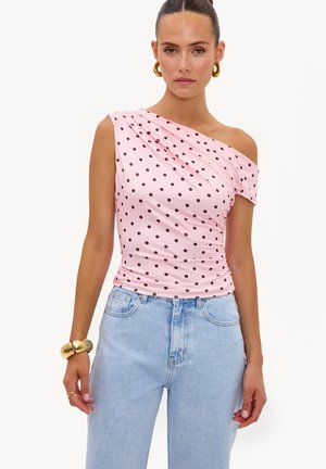 Femme portant un top rose asymétrique à pois noirs, un jean taille haute bleu clair, des boucles d’oreilles créoles dorées et un bracelet doré épais.