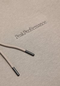 Beige hættetrøje med en struktureret stof, der har den broderede tekst "Peak Performance" og kontrasterende snore med metalspidser.