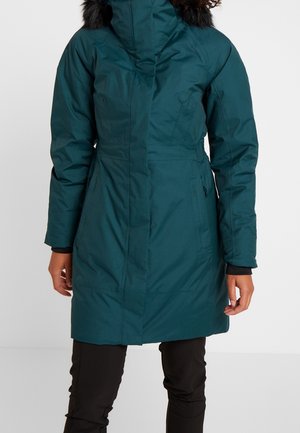 Osoba v tyrkysovém zimním kabátu s kapucí lemovanou černým kožíškem a v černých kalhotách stojí na jednobarevném světle šedém pozadí.