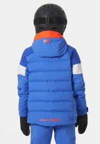 Modrá péřová bunda s černými rukavicemi, která má oranžovo-bílé barevné blokování. Logo je na zádech a kapuci. Hladký, izolační materiál.