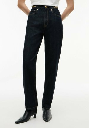 Kvinde iført højtaljede mørkeblå jeans, hvid top stoppet ind, sorte spidse ankelstøvler, stående med hænderne bag ryggen.