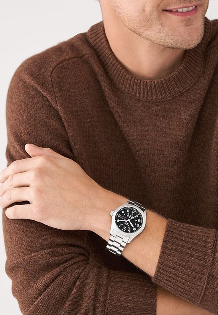 Reloj plateado con esfera negra, que presenta números blancos en negrita y función de fecha. Lleva puesto en una muñeca sobre un suéter de punto marrón.