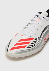 Zapatilla deportiva blanca con rayas rojas y negras, cordones blancos y acentos plateados, mostrando los detalles de la punta y el lado sobre un fondo blanco.