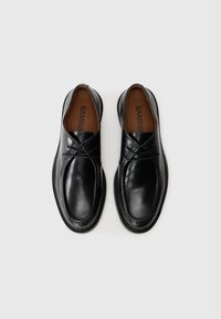 Black leather shoes with a rounded shape, featuring cross-over detailing, smooth texture, and a stitched sole. Brown inner lining visible.