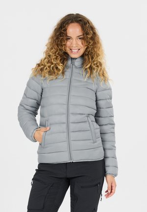 Frau mit lockigem Haar, die eine graue gesteppte Jacke und schwarze Hose trägt, die Hände in die Taschen gesteckt hat und vor einem einfarbigen Hintergrund lächelt.