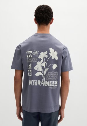 Mężczyzna noszący szarą koszulkę z białymi botanicznymi i pszczelimi ilustracjami oraz tekstem o naturze i zrównoważonym rozwoju na plecach.