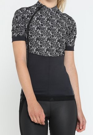 Nainen pukeutuneena istuvaan musta-harmaaseen kukkakuviolliseen lyhythihaiseen toppiin ja mustiin housuihin, seisoo käsivarret rentoina sivuilla.