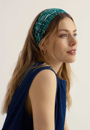 Fiatal nő kék ujjatlan felsőben, zöld mintás fejpánttal, oldalra néz enyhe mosollyal, egyszínű háttér előtt.