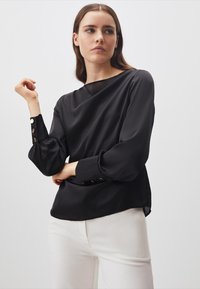 Blusa di satin nera con scollatura a barca, maniche a sbuffo e bottoni decorativi sui polsini. Abbinata a pantaloni bianchi per un contrasto.
