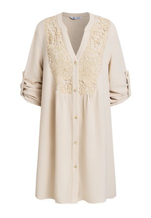Chemise beige boutonnée avec un motif floral en dentelle sur la poitrine. Présente des manches longues et un tissu texturé.