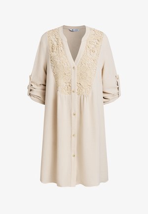 Beige overhemd met knopen en een kanten bloemenpatroon op de borst. Heeft lange mouwen en een structuurstof.