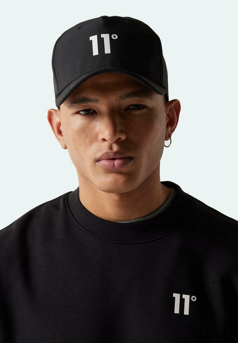 Gorra negra con el logotipo blanco "11°", llevada por una persona en un sudadera negra. Incluye un visera curva y una correa trasera ajustable.