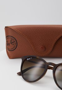 Óculos de sol Ray-Ban com armação em tartaruga castanha e lentes degradê, ao lado de uma caixa de couro texturizado castanho com logótipo em relevo.