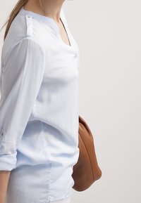 Blusa a maniche lunghe di colore azzurro chiaro con scollatura a V, dettagli in bottone sulle spalle e una texture liscia e morbida. Abbinata a una clutch in pelle marrone.