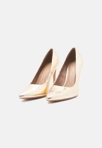 Um par de sapatos de salto alto dourados e brilhantes para mulheres, com bicos pontudos, num fundo branco.