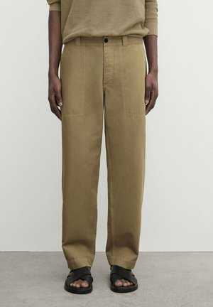 Person trägt lockere khakifarbene Hose, beige gerippten Pullover und schwarze Sandalen mit offenen Zehen, steht auf grauem Boden vor weißer Wand.