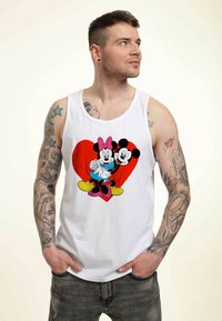 Camiseta sin mangas blanca con un gráfico colorido de Mickey y Minnie Mouse frente a un corazón rojo. El modelo lleva pantalones vaqueros grises, mostrando tatuajes en los brazos.