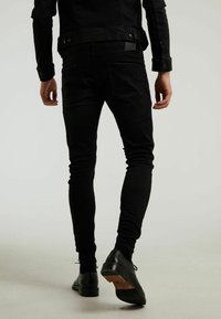Jeans de mezclilla negra con corte ajustado. Presentan una textura suave y bolsillos traseros, combinados con zapatos de vestir negros y una chaqueta negra.