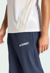 Weißes Sportshirt mit Netzpaneelen und hellen Streifen, kombiniert mit marineblauen Hosen mit prominentem weißem "TERREX"-Logo.