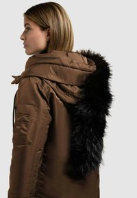 Parka marrón con capucha forrada, que presenta un ribete de piel sintética negra y un bolsillo con cremallera en la manga. Textura suave y brillante.