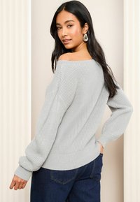 Pulover tricotat gri deschis cu un design off-the-shoulder, mâneci lungi, tivuri ondulate și o croială lejeră, asortat cu blugi de culoare albastru închis.