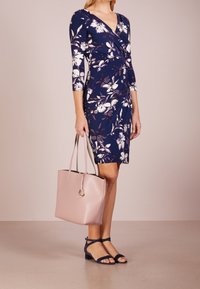 Marinblå blommig omslagsklänning med trekvartslånga ärmar, i kombination med en mjuk rosa tote-väska och blå snörsandaler på en texturerad yta.