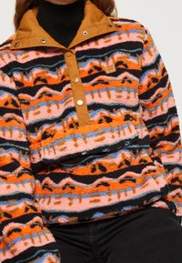 Bluza z mikrofibry z pomarańczowo-różowym wzorem zygzakowym, czarnymi akcentami i kołnierzem na guziki, wykonana z miękkiego, teksturowanego materiału.