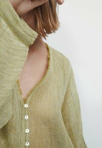 Lichtgroene, getextureerde blouse met V-hals, met een raw edge en witte knopen in het midden. Zacht, vloeiend materiaal.