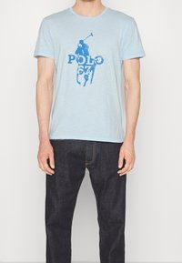 Světle modré bavlněné tričko s grafikou modrého jezdeckého hráče a textem "POLO 67". Krátké rukávy a kulatý výstřih.