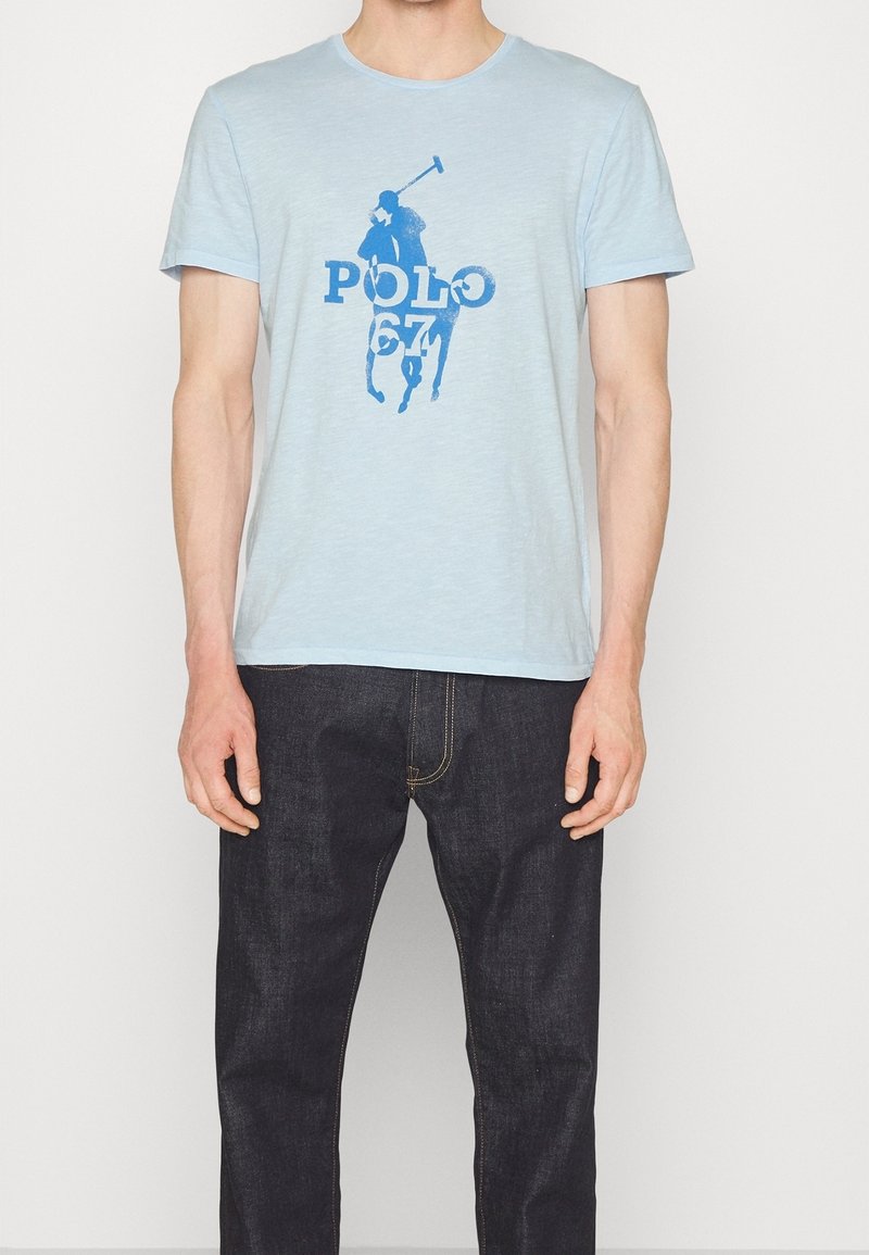 Světle modré bavlněné tričko s grafikou modrého jezdeckého hráče a textem "POLO 67". Krátké rukávy a kulatý výstřih.